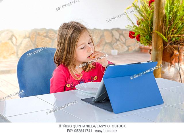 Four years old blonde girl with red shirt sitting watching digital tablet and eating biscuits on the table