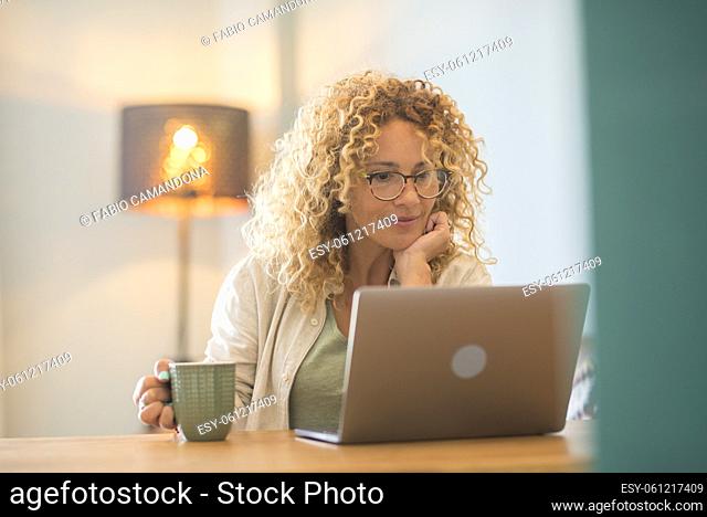 Beautiful caucasian young woman with curly hair and eyeglasses drinking coffee and looking at laptop screen. Woman drinking coffee while using laptop at home...