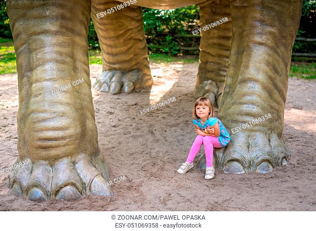 Little girl sitting under huge life sized Diplodocus dinosaur sculpture in an amusement park in summer