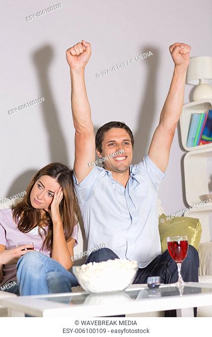 Excited man watching TV with wine and popcorn by bored woman at home