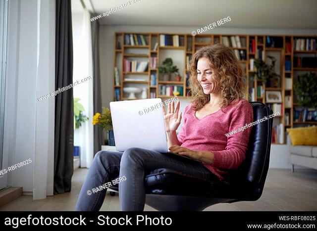Smiling woman waving hand to video call on laptop while sitting at home