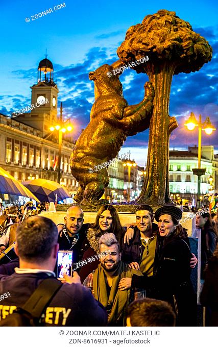 Madrid, Spain - Jan 23, 2016: Group of friends posing by statue of a bear and a madrone tree on busy Puerta del Sol square at dusk on January 23th