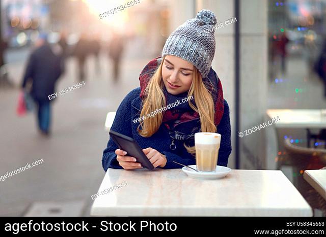 Young woman sitting enjoying a drink outdoors in winter at a street cafe sitting holding her mobile phone or small tablet looking at the camera with a friendly...
