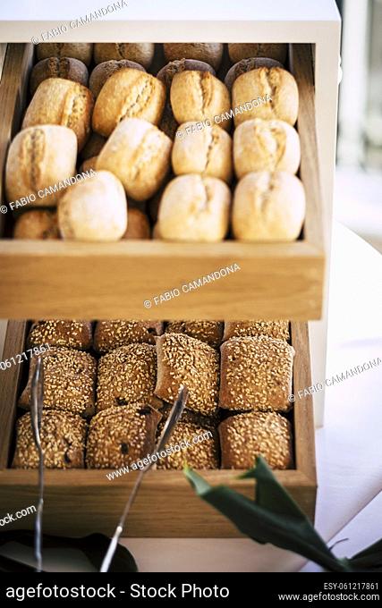 Bread buns in abundance on display at slatted shelf fixture in hotel or supermarket for sale. Freshly baked bread buns displayed in a row on wooden shelf with...