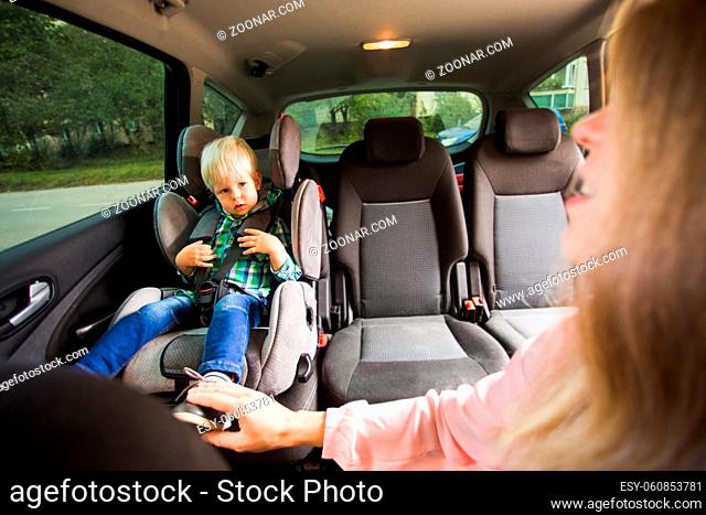 The happy mom is sitting behind the wheel of a car and looks back at her baby. The mom touches her baby boy who is sitting at the back of a car seat
