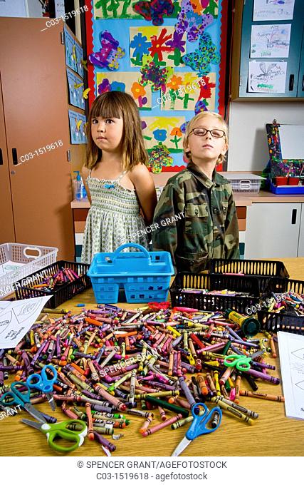 Kindergarten children in San Clemente, CA, contemplate a table full of classroom objects including crayons and worksheets