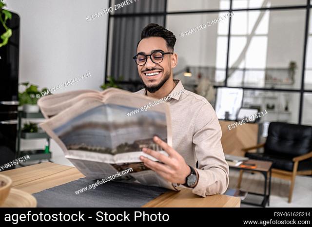 Life style. Happy attractive successful man in glasses smiling reading newspaper at home sitting at table