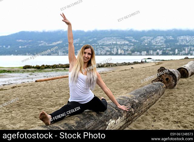 Young woman at the sandy beach of Stanley Park stands barefoot on log, her arms are raised and doing some acrobatic exercise movement, Vancouver