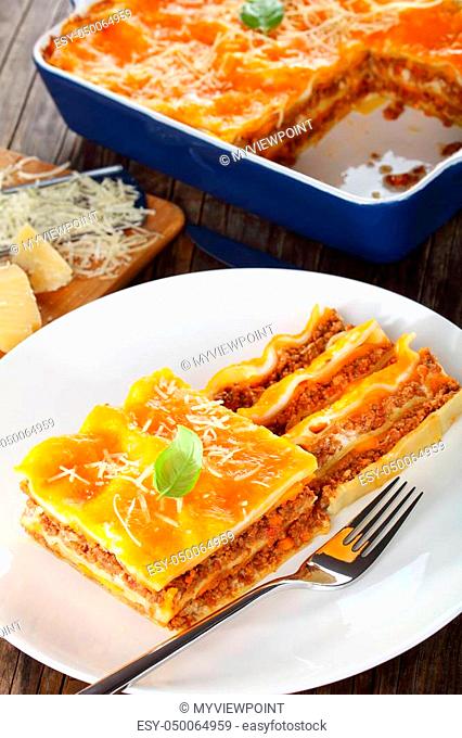 portion of italian lasagna topped with melted cheese and fresh basil leaves, sprinkled with grated parmesan cheese on white plate with fork, authentic recipe