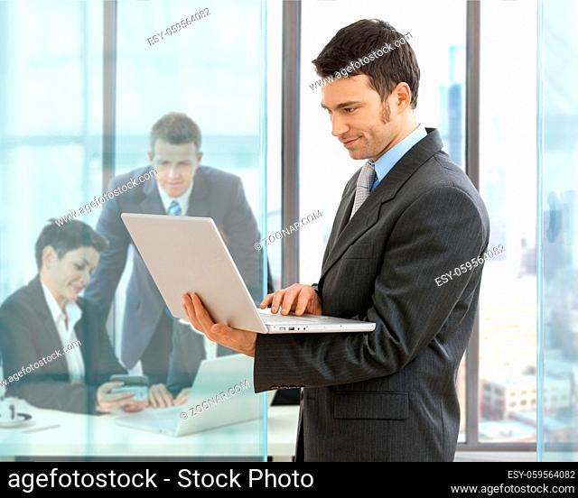 Businessman using laptop standing in office, businesspeople working at desk in the background