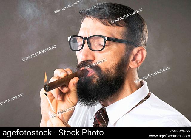 The elegant bearded man with eyeglasses is lighting a cigar on gray background
