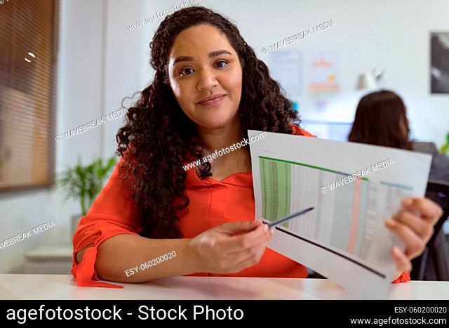 Smiling biracial businesswoman sitting at desk making video call holding documents in modern office