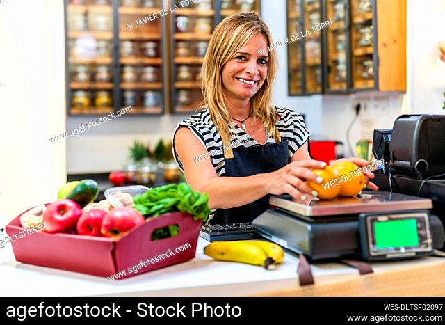 Smiling saleswoman weighing oranges on weight scale at checkout counter in store
