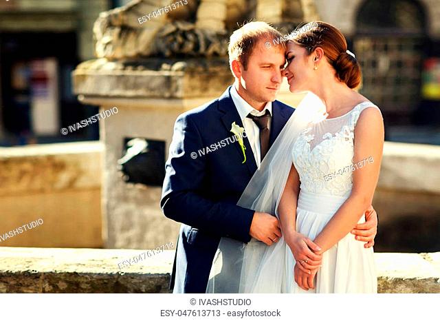 A peaceful groom holds a tender bride in his arms while they stand behind an old fountain