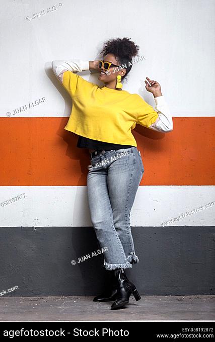 Full body of confident positive young African American female wearing trendy jeans and bright yellow shirt with sunglasses and earrings leaning against colorful...