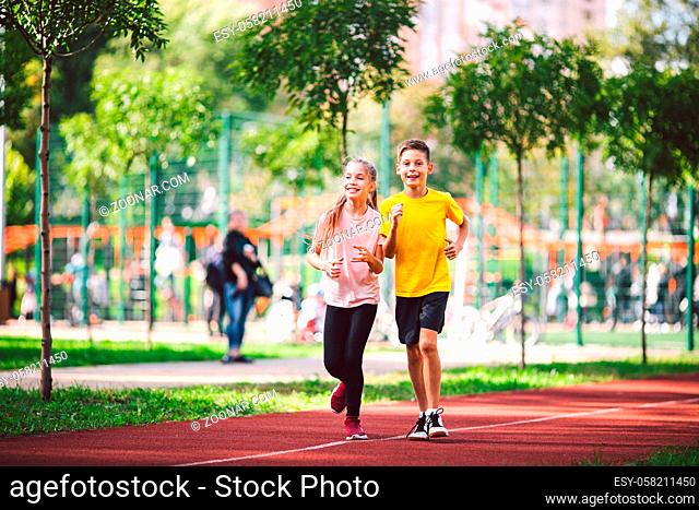 Couple of kids boy and girl doing cardio workout, jogging in park on jogging track red. Cute twins runing together. Run children, young athletes