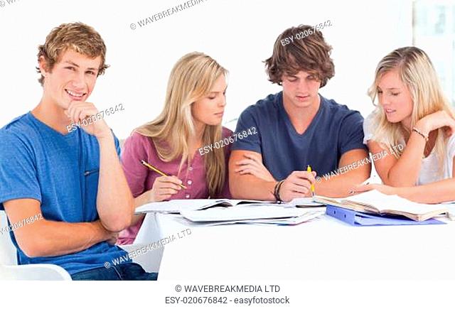 A group of students sitting together as they all study as one smiles