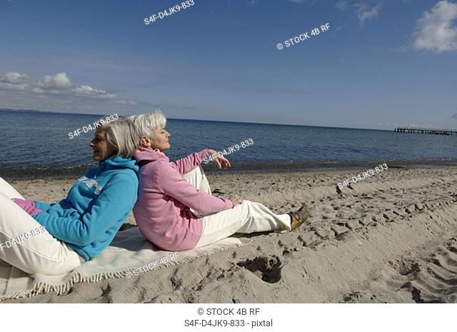 Two mature women sitting back to back at Baltic Sea beach
