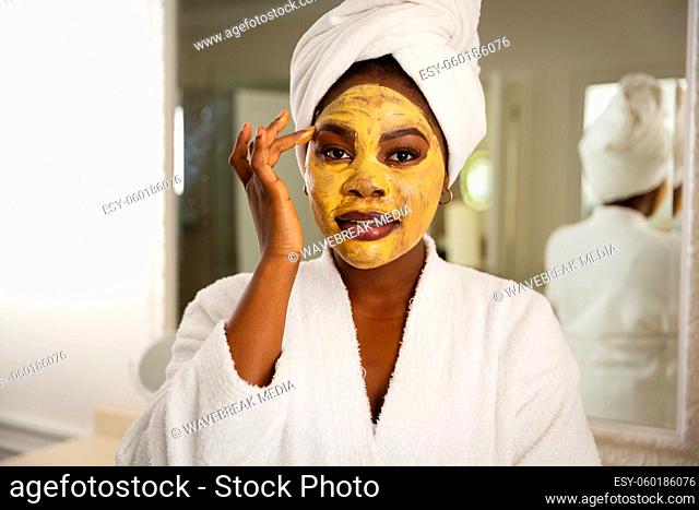 Happy african american woman in bathroom with towel on head, looking in mirror applying face mask