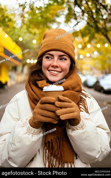 Smiling woman in warm clothing holding disposable coffee cup on sidewalk