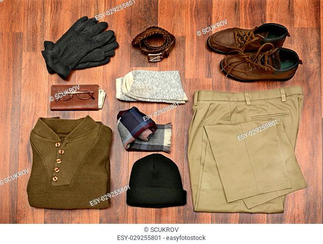 High angle shot of men's winter clothes laid out on a dark wood floor. Items include, Sweater, Scarf, Gloves, wool Socks, Pants, Boots, belt, Knit Cap, Wallet