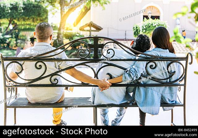 Unfaithful woman sitting holding hands with another man while boyfriend hugs her. Unfaithful couple holding hands on a park bench