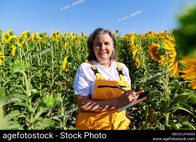 Smiling female farm worker holding book while standing amidst sunflowers