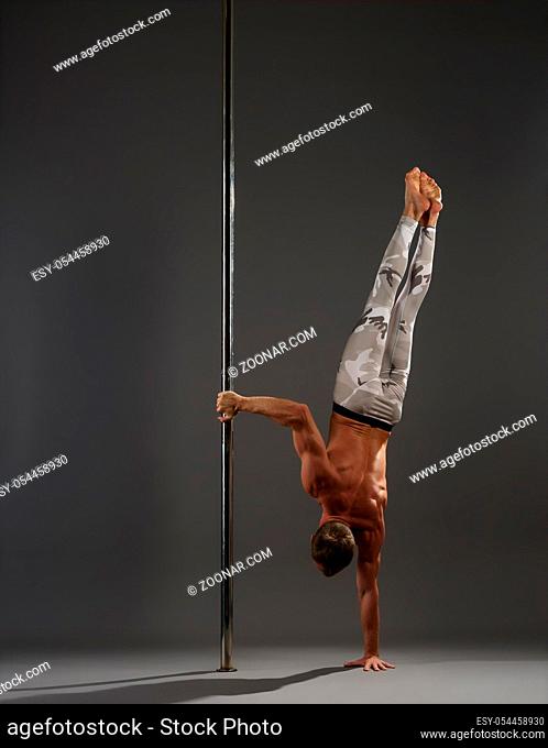 Athletic young pole dancer in leggings standing on one hand by pylon in studio