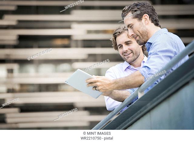 Two men on an urban walkway, looking at a digital tablet