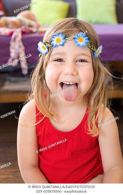 portrait of three years old blonde pretty girl face looking and sticking out tongue, with red sleeveless t-shirt and wreath with flowers, indoor at home