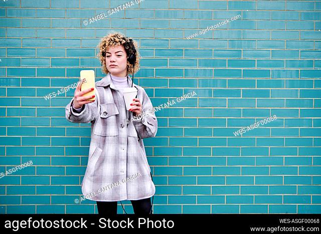 Woman with headphones holding disposable coffee cup while taking selfie through mobile phone against blue wall