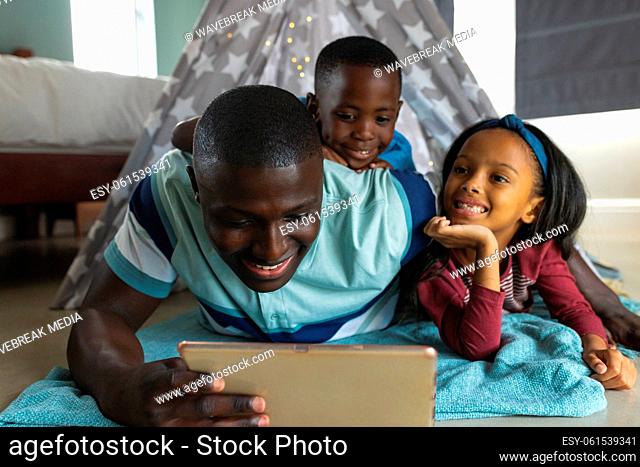 Smiling multiracial father using digital tablet while relaxing with children in blanket tent at home