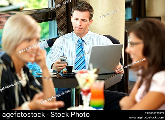 Businessman using laptop and mobile in cafe, young woman eating sweets and talking in the foreground. Selective focus on businessman