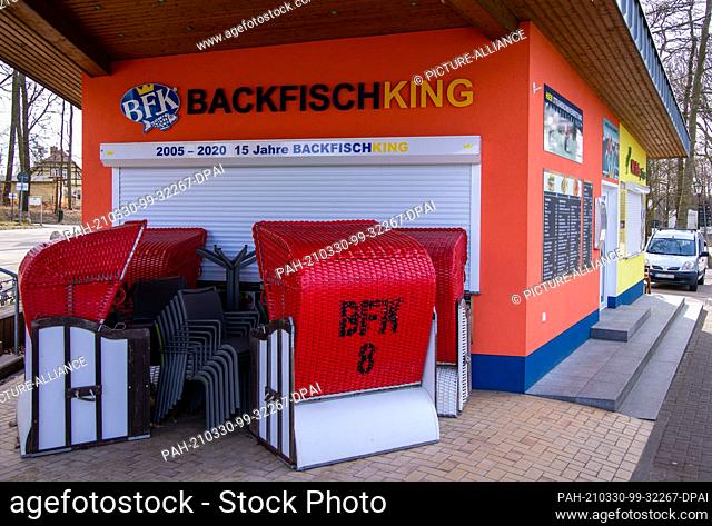 22 March 2021, Mecklenburg-Western Pomerania, Koserow: Beach chairs and terrace chairs stand in front of the closed sales stand for baked fish on the beach near...