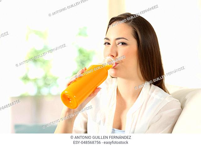 Woman drinking orange juice from a bottle looking at you sitting on a couch in the living room at home