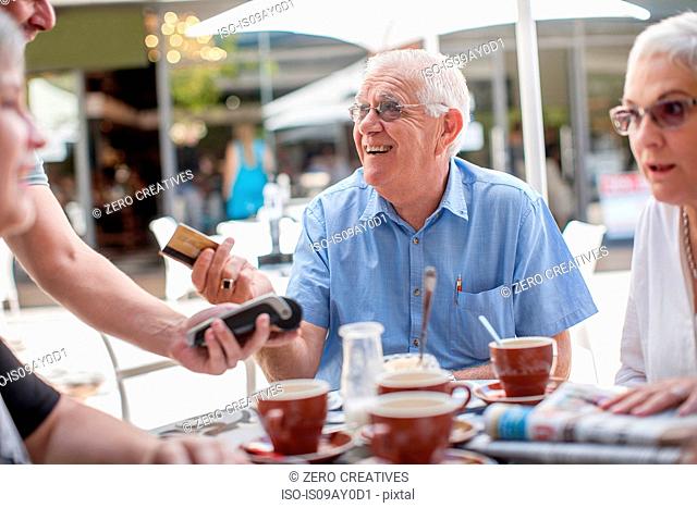 Group of mature friends, sitting outside cafe, senior man handing credit card to waiter