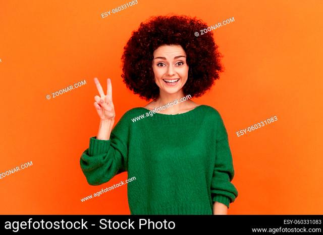 Happy friendly woman with Afro hairstyle wearing green casual style sweater looking at camera with toothy smile, showing v sign, peace gesture