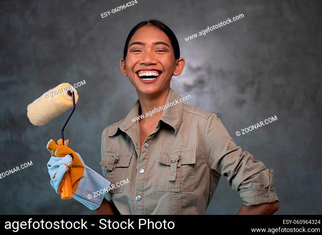 Portrait of attractive young girl builder with paint roller in overall and gloves smiling over concrete wall background