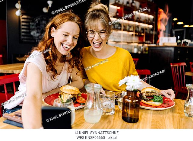 Two happy female friends having burger and taking a selfie in a restaurant