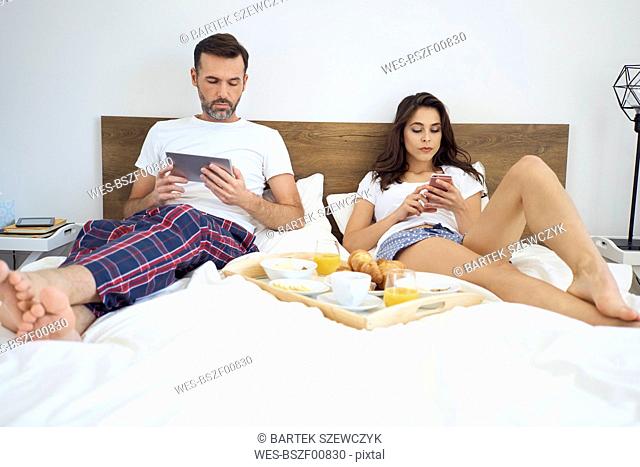 Bored couple lying in bed with breakfast, using mobile devices