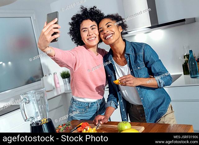 Happy friends taking selfie though smart phone standing in kitchen at home