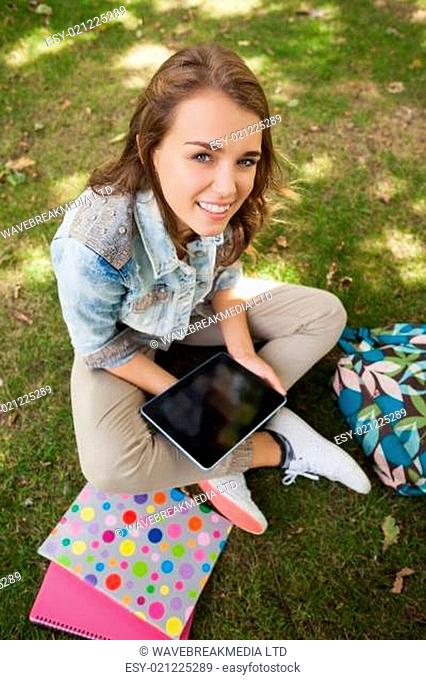 Pretty cheerful student sitting on grass using tablet