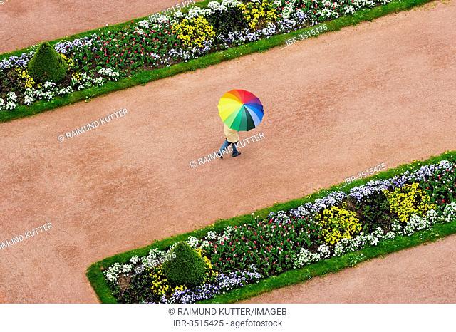 Person with an umbrella in rainbow colours on a red dirt road between flowerbeds in the Palace Gardens, Stadtschloss City Palace