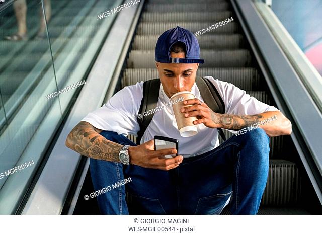 Young man with backpack sitting on escalator looking at cell phone while drinking coffee to go