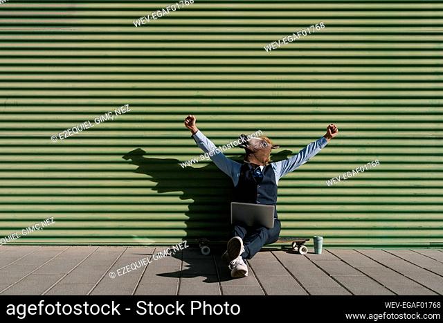 Businessman in horse mask with laptop gesturing while sitting on longboard against wall
