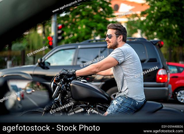 Smiling man on the black motorcycle stands in the traffic before the red traffic light. He wears blue ripped jeans, a gray T-shirt