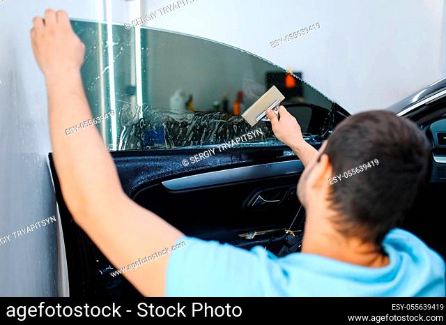 Male worker with squeegee installs car tinting, tuning service. Mechanic applying vinyl tint on vehicle window in garage, tinted automobile glass