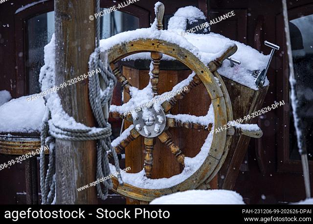 06 December 2023, Mecklenburg-Western Pomerania, Kirchdorf (poel): The wooden steering wheel of a historic sailing ship is covered in snow in the harbor on the...