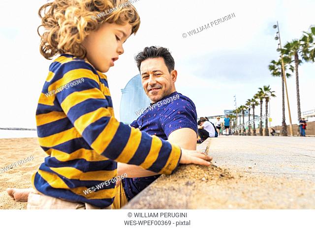 Spain, Barcelona, young boy playing with sand, his father sitting next to him and smiling