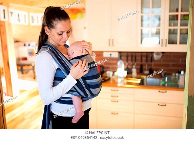Beautiful young mother in kitchen with her baby son sleeping in sling at home, gently stroking him